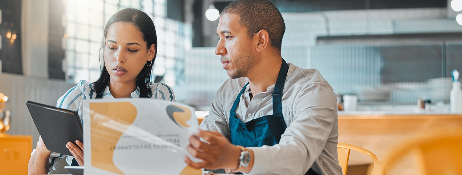 Two business owners in the restaurant industry reviewing their budget and business plans while sitting on a brightly lit empty restaurant room