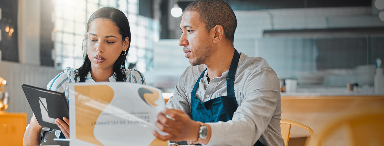 Two business owners in the restaurant industry reviewing their budget and business plans while sitting on a brightly lit empty restaurant room