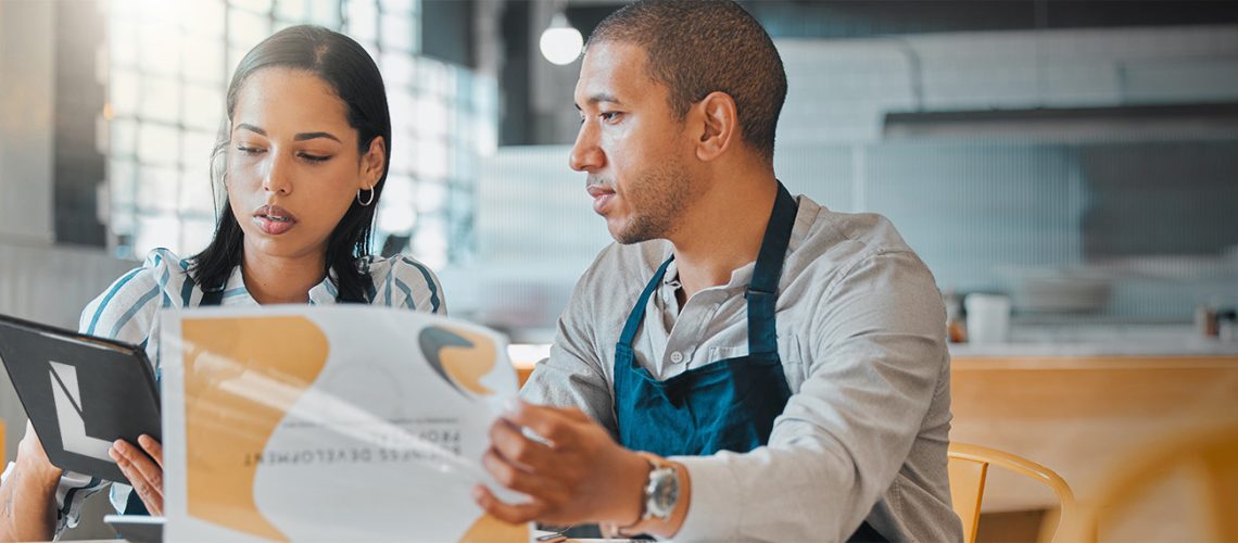 Two business owners in the restaurant industry reviewing their budget and business plans while sitting on a brightly lit empty restaurant room