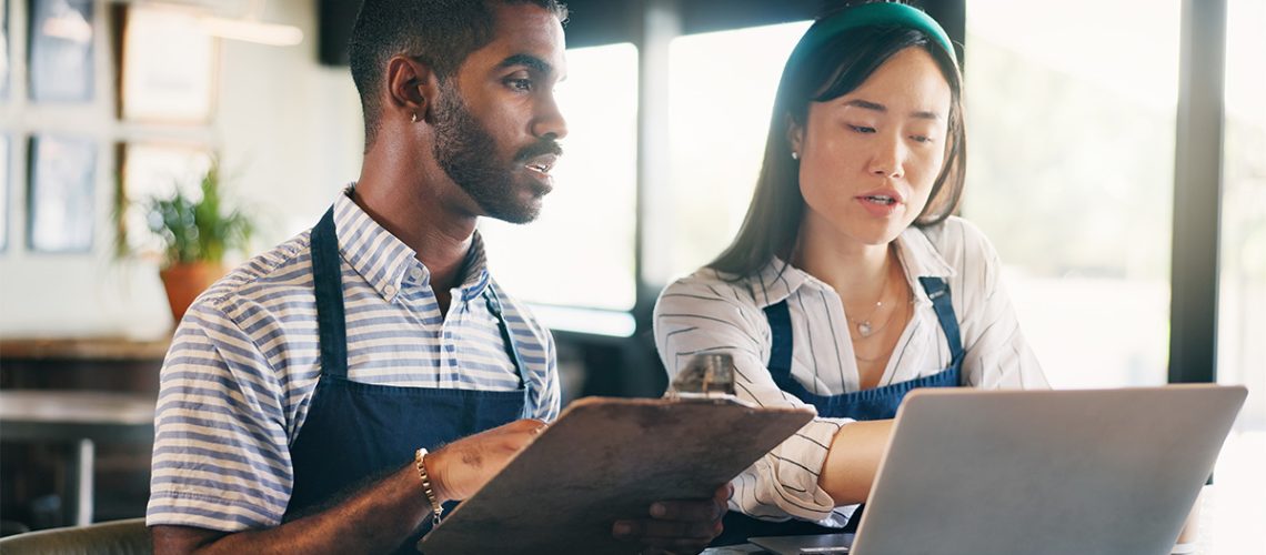 Two business owners in the restaurant industry reviewing their budget and business plans while sitting on a brightly lit empty restaurant room