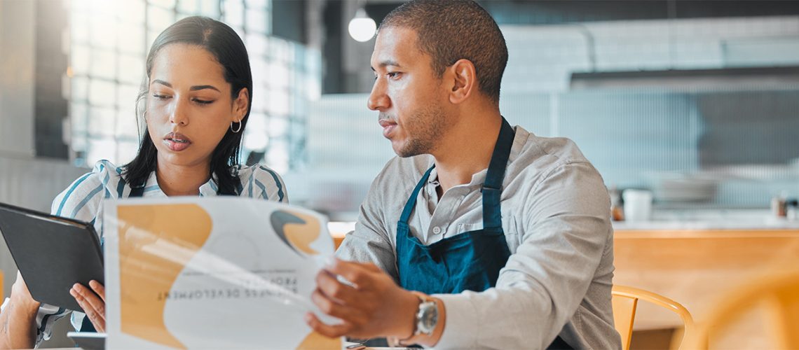 Two business owners in the restaurant industry reviewing their budget and business plans while sitting on a brightly lit empty restaurant room